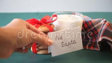 圣诞老人的圣诞饼干。 女人`手在牛奶、饼干、松枝和餐巾纸的玻璃旁边放了一条短信给圣诞老人。